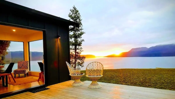 Un área de estar moderna al aire libre con dos sillas y una mesa, con vistas panorámicas a un lago y montañas durante el atardecer.