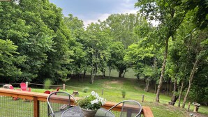 A deck with metal chairs and a table, surrounded by a wooden railing, overlooking a grassy area with trees.