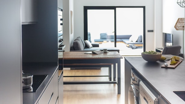 A modern kitchen with a central island, a dining area with a sofa and table, and a large window offering a view of the outdoors.
