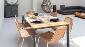 A dining area with a table set for two, surrounded by wicker chairs, a large wall clock, and a modern sofa.