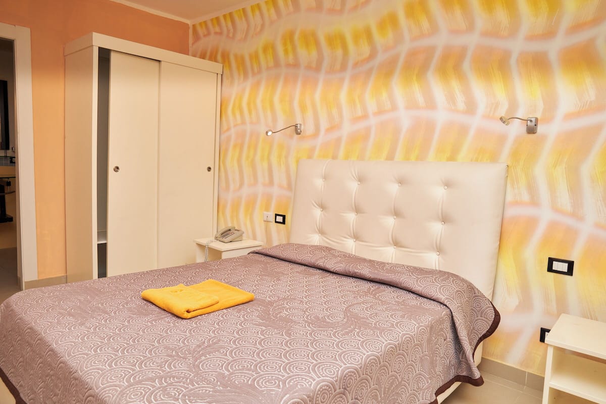 Een slaapkamer met een gestuukt hoofdeinde, een bed met een dessin, een nachtkastje met een lamp, en een wand met een geometrisch patroon.
