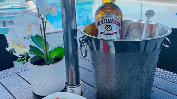 Escena junto a la piscina con una botella de Chivas Regal, un plato de sandía en rodajas y una orquídea en maceta.