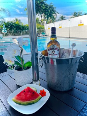 Un cadre agréable au bord de la piscine, avec une bouteille de Chivas Regal, une assiette de tranches de pastèque et une orchidée en pot.