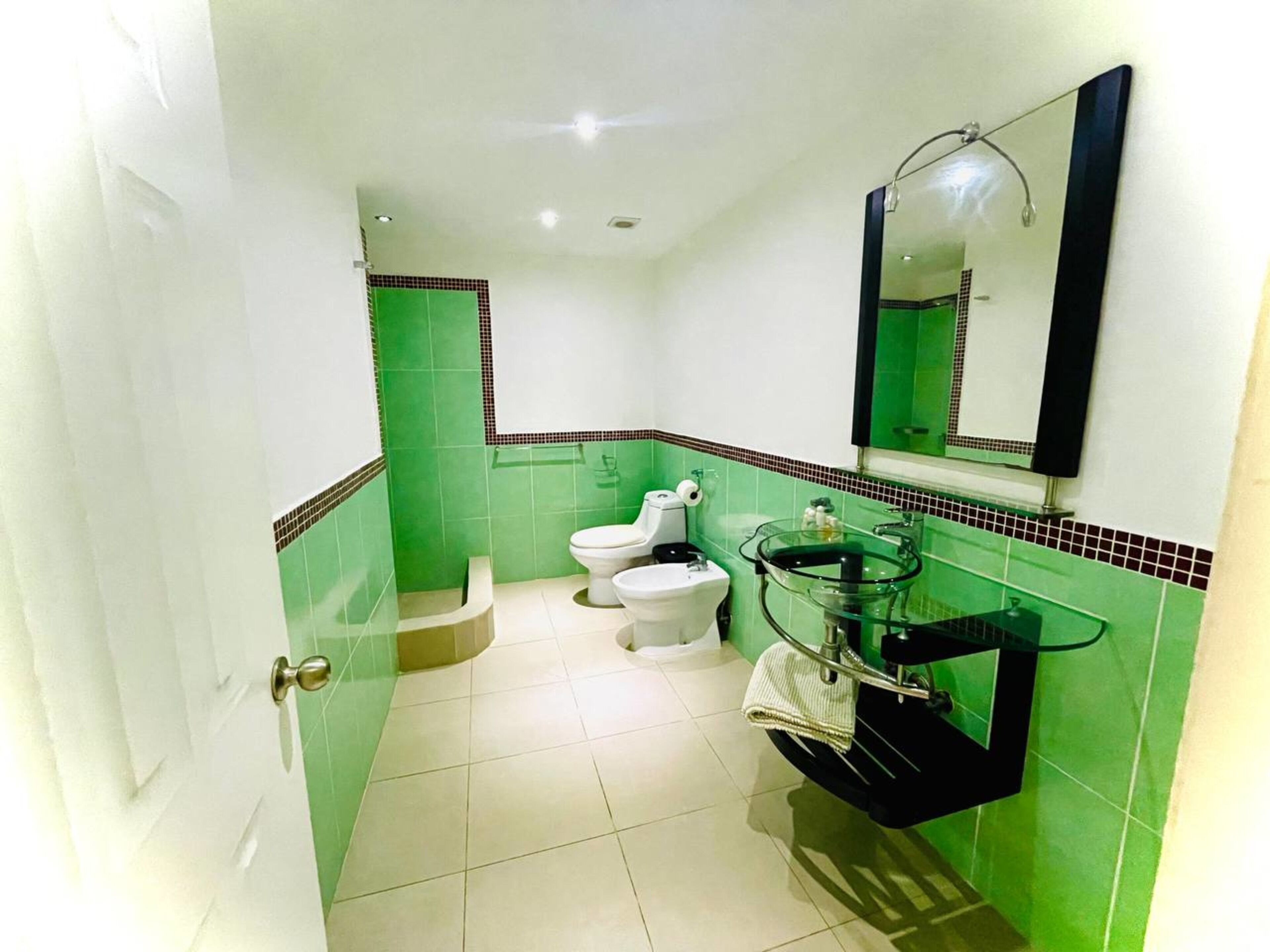 A bathroom with green tiles, a glass sink, a toilet, and a mirror.