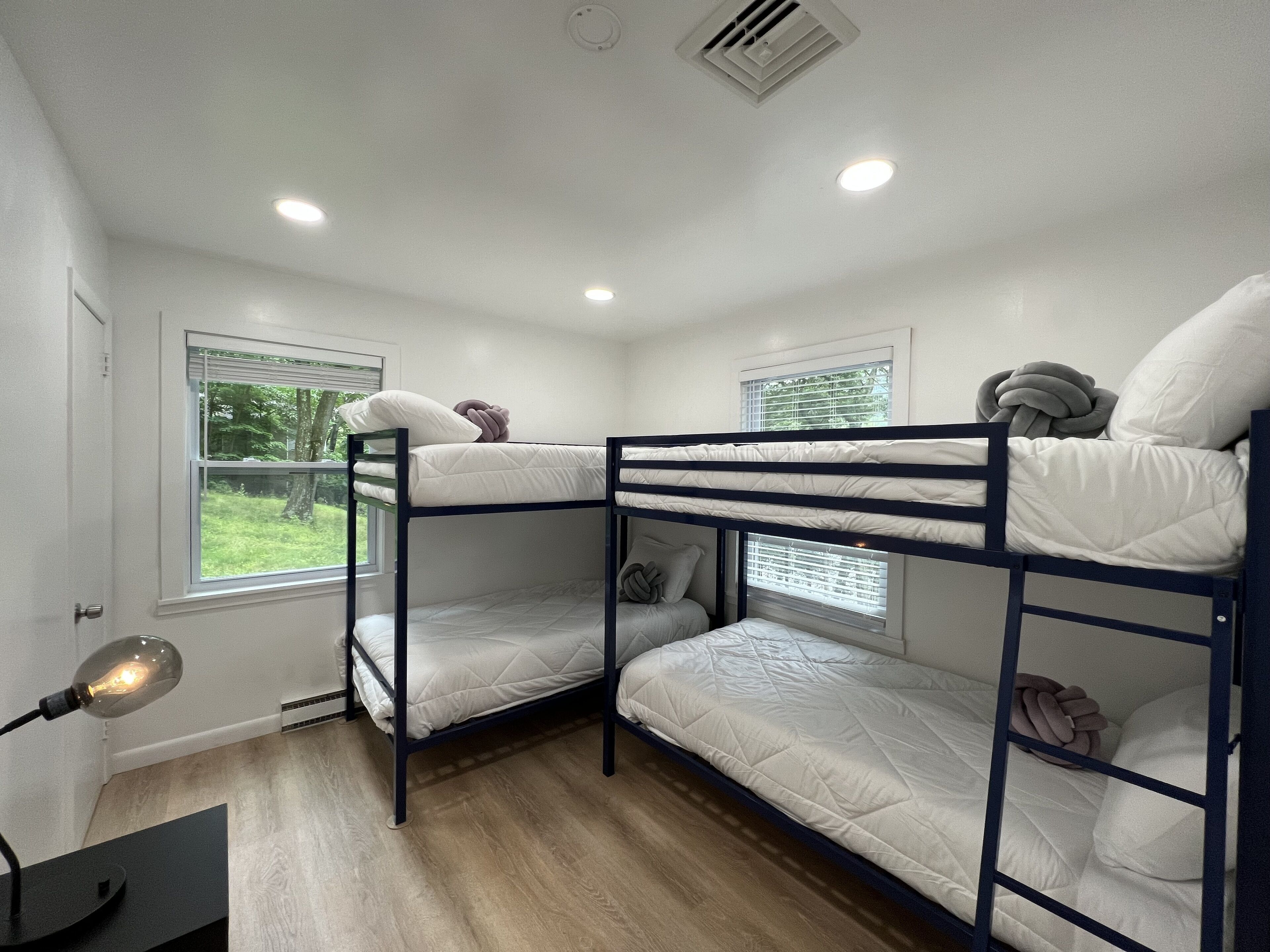 Une chambre avec un lit superposé, deux lits, un bureau avec une lampe et deux fenêtres équipées de stores.