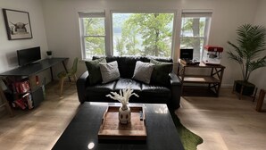 A living room with a black leather couch, a wooden coffee table, and a TV on a stand. There are three windows with a view of trees and a lake.
