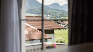 Blick durch ein Fenster auf ein mit Ziegeln gedecktes Dach, eine Kirche mit einem Glockenturm und Berge im Hintergrund.