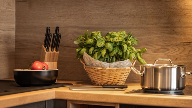 Una encimera de cocina con una cesta tejida que contiene albahaca, una olla y un bol con tomates.