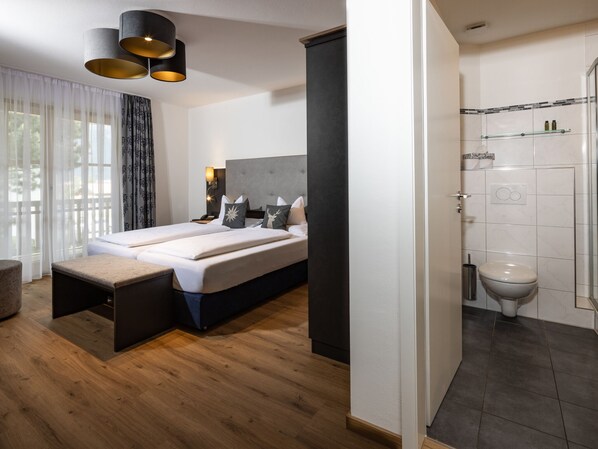 A modern hotel room with a large bed, bedside tables, a nightstand, a wall-mounted lamp, and a view of the outdoors through sheer curtains.