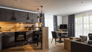 Una cocina moderna con armarios oscuros, encimera de madera y horno empotrado. Hay una zona de comedor con una mesa y sillas, y una sala de estar con sofá y una mesa de centro.