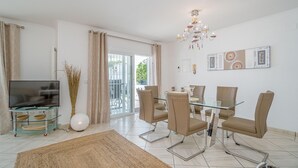Una sala de estar moderna con una mesa de comedor de vidrio, sillas color beige, un televisor de pantalla plana sobre un pedestal de vidrio y una araña.
