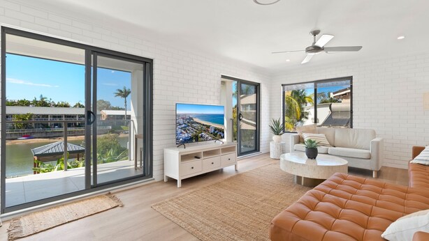 Una sala de estar moderna con ventanas amplias, un televisor de pantalla plana, un ventilador de techo y vistas a una playa y edificios.