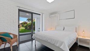 Un dormitorio moderno con una cama grande, una silla de mimbre, una puerta corrediza de vidrio a un balcón y una mesita de noche con una lámpara.