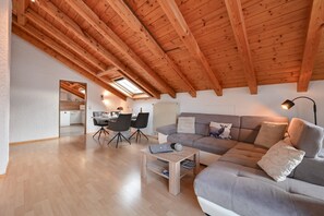 A modern loft apartment with a high wooden ceiling, a cozy living area with a sofa and coffee table, a dining area with chairs, and a kitchen in the background.