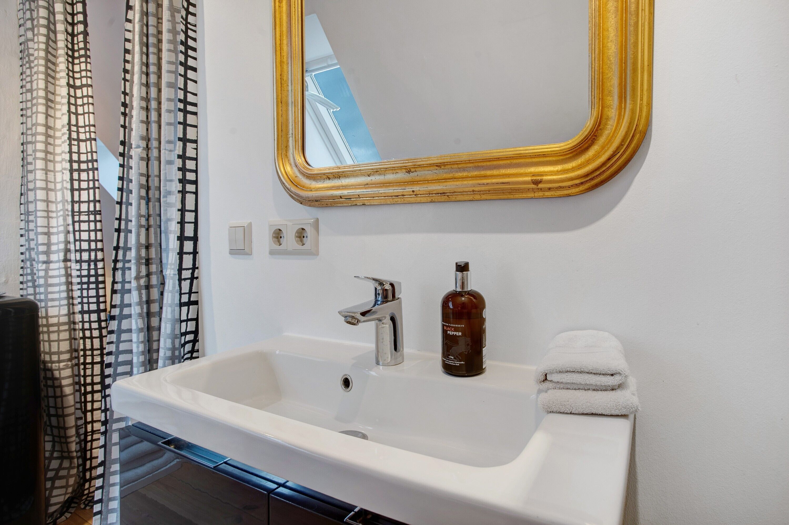 Ein Badezimmer mit einem weißen Waschbecken, einem spiegel mit goldener Rahmen, einer Seife und einem Handtuch.