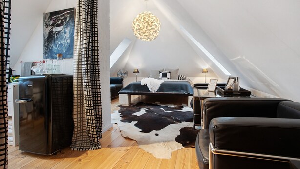 A bedroom with a bed, a black leather sofa, a cowhide rug, a refrigerator, a lamp, and a ceiling fan.