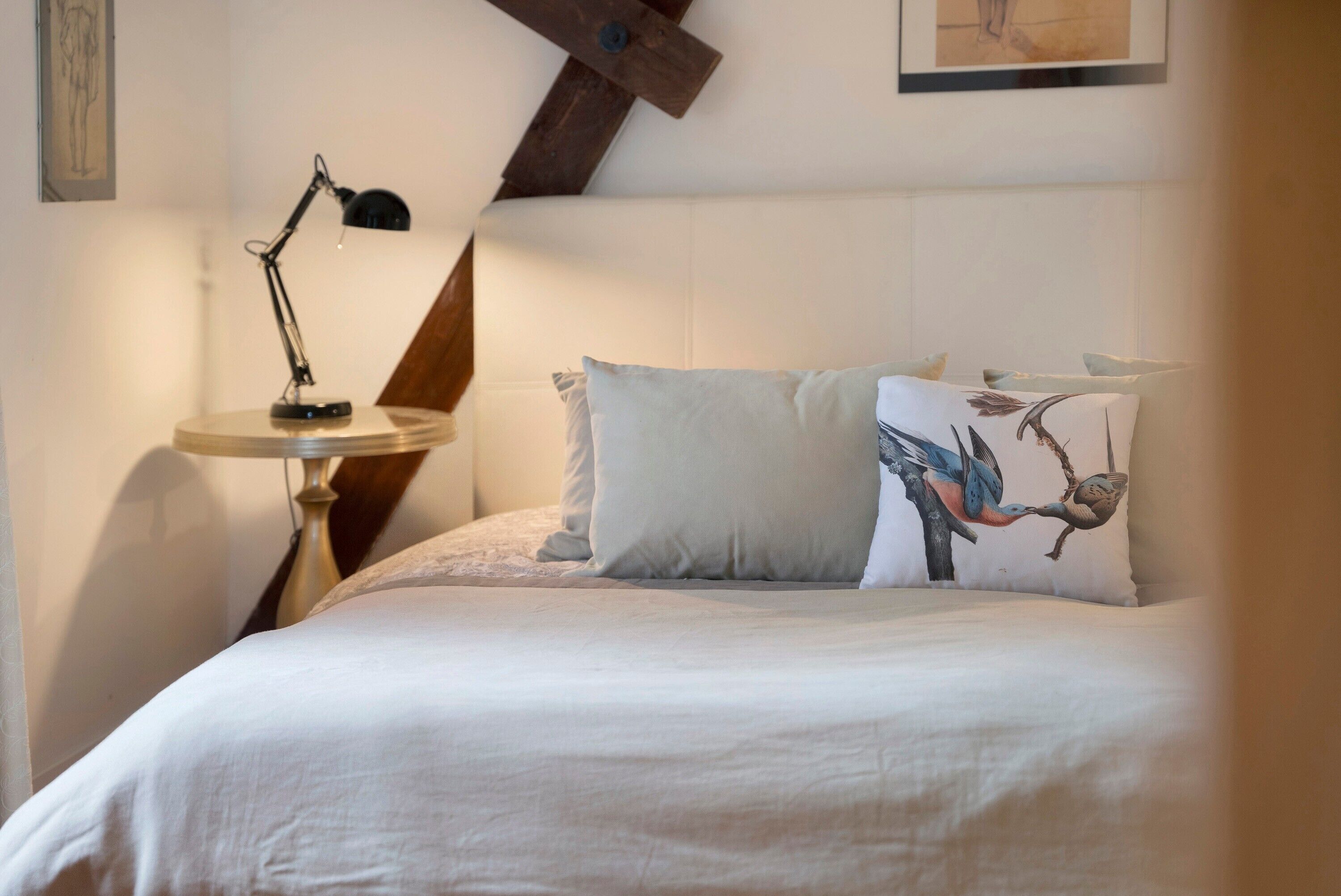 Een netjes opgemaakt bed met een sierkussen met een vogelmotief, een klein rond tafeltje met een lamp en een houten balk in het plafond.