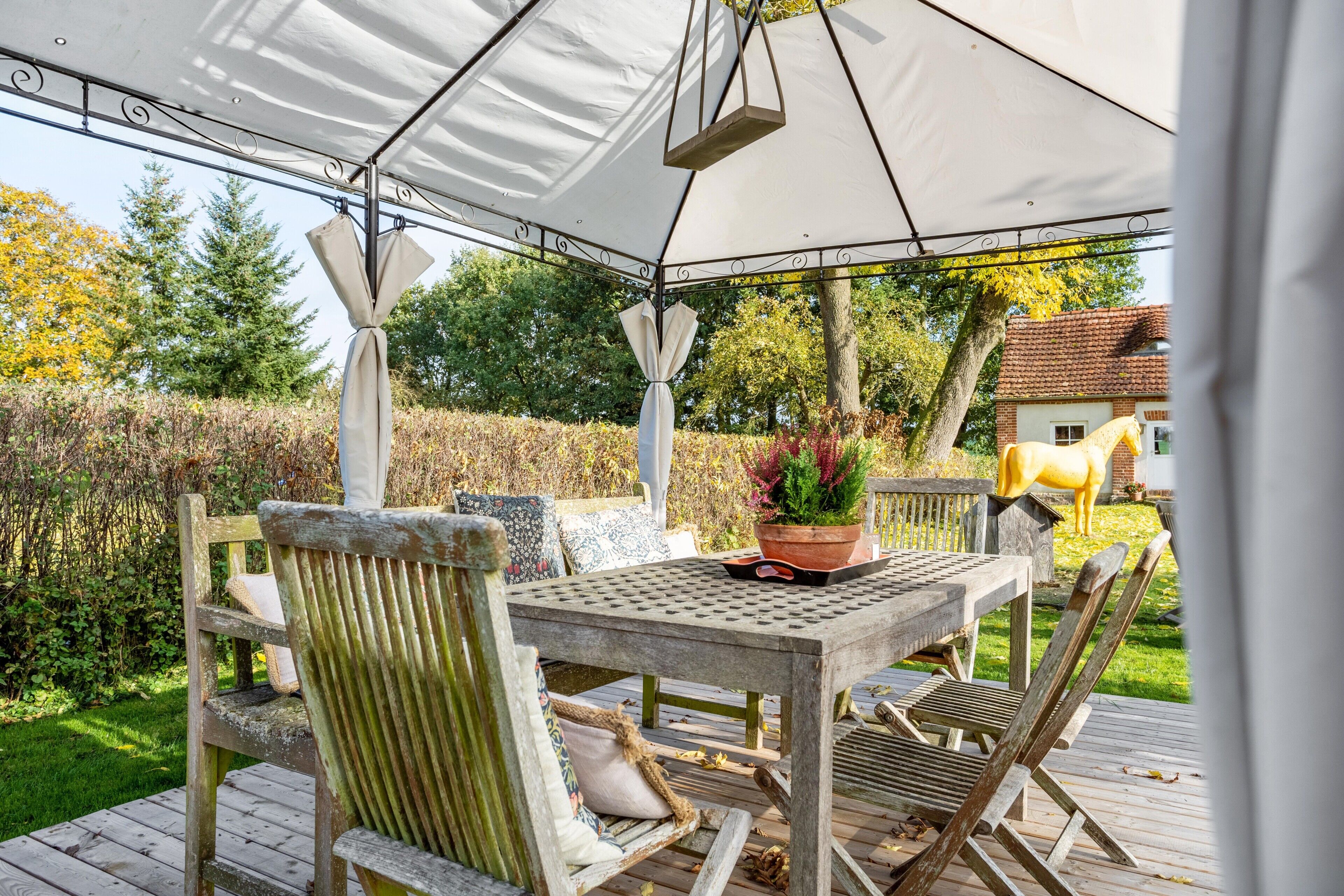 A covered outdoor seating area with a table and chairs, surrounded by a garden and a house in the background.