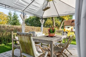 A covered outdoor seating area with a table and chairs, surrounded by a garden and a house in the background.