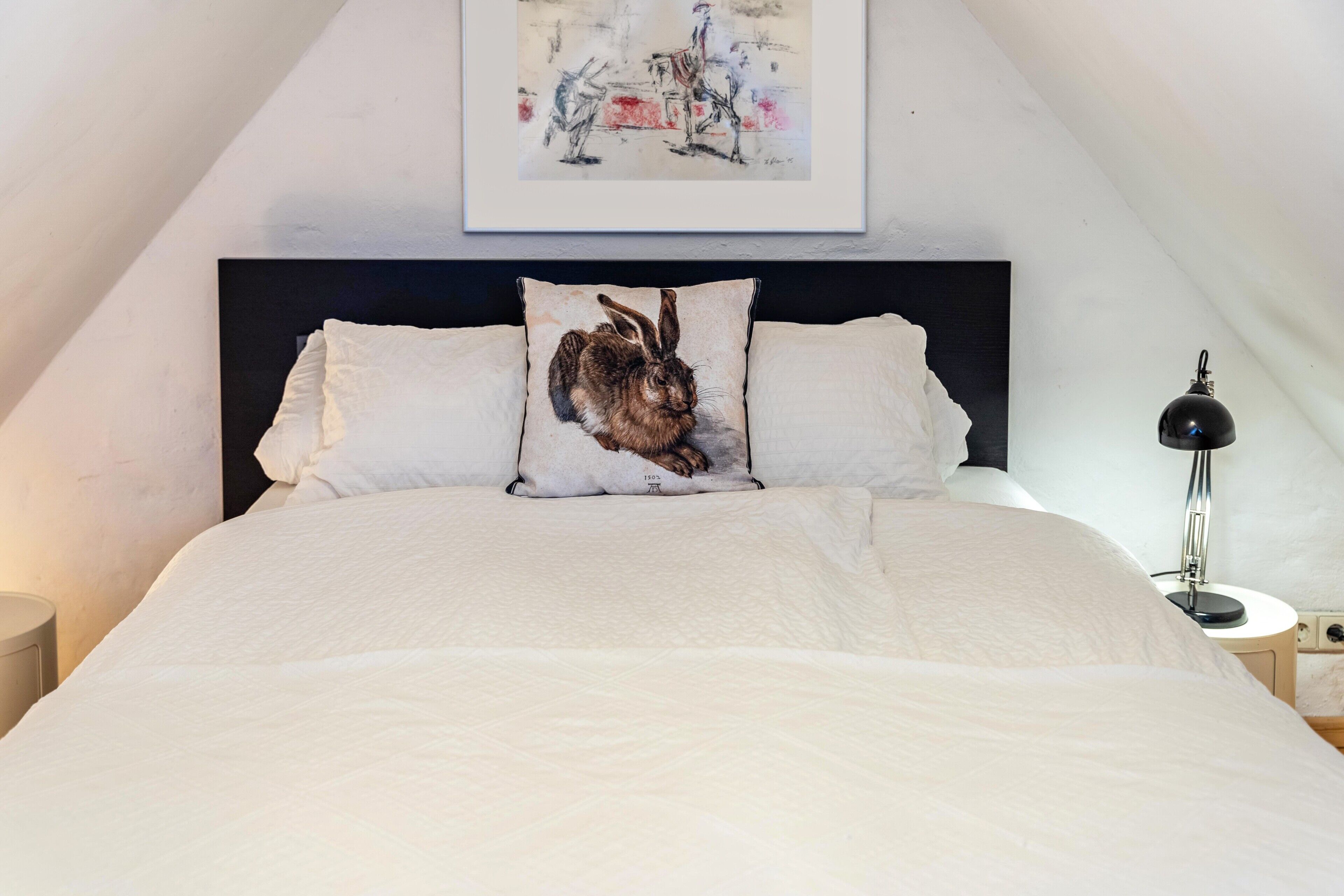 Een netjes opgemaakt bed met witte beddengoed, een kussen met een konijntjesprint, een zwart hoofdbord, een nachtkastje met een lamp en een lijst met een kunstwerk erboven.