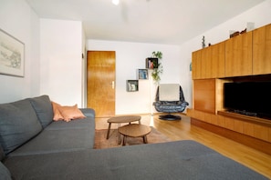 Un salón con un sofá gris, una mesita de madera, una silla azul, una estantería y un televisor.