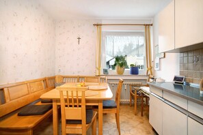 Un comedor con una mesa y bancos de madera, una ventana con cortinas, un radiador y una zona de cocina con armarios y una encimera.