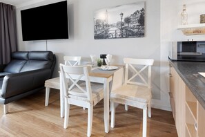 Una sala moderna con un sofá de cuero negro, una mesa de comedor con sillas blancas, un televisor de pantalla plana montado en la pared y una pintura de un paisaje urbano en blanco y negro sobre la mesa.