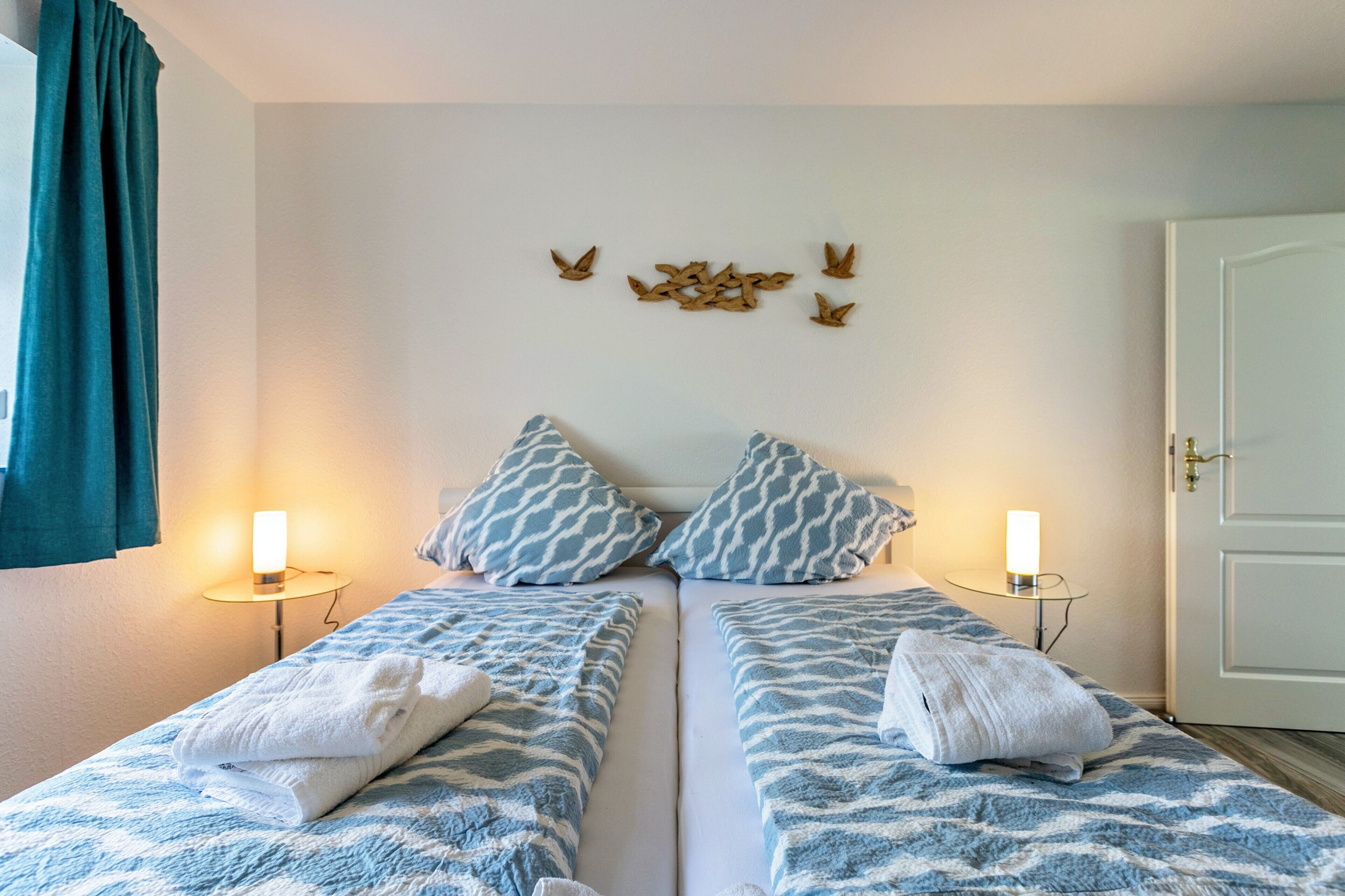 Habitación doble con dos camas, cada una con una colcha estampada y almohadas a juego. Hay luces fijadas a la pared a ambos lados de las camas, y una puerta a la derecha.