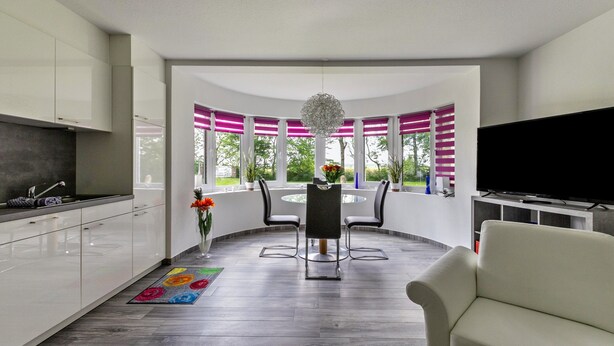 A modern kitchen with white cabinets, a dining area with a round table and chairs, a TV, and a view of greenery through large windows.