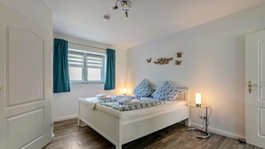 A bedroom with a bed, a window with curtains, a bedside table with a lamp, and a wall-mounted light fixture.