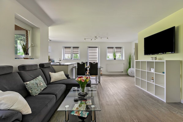 Un salon moderne avec une table basse en verre, un canapé agrémenté de coussins, un téléviseur à écran plat fixé au mur, et un coin cuisine à l’arrière-plan.