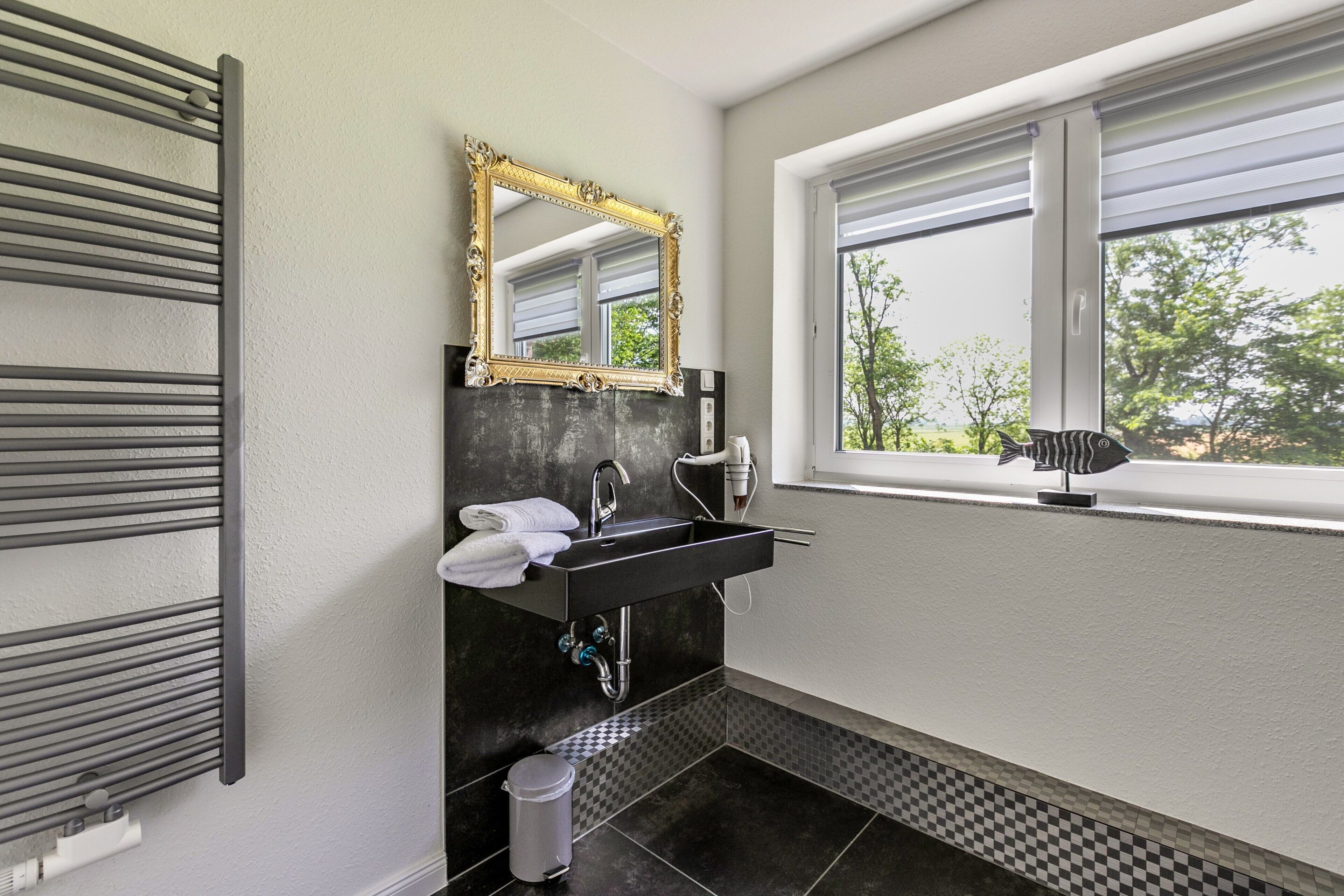 Une salle de bain moderne avec un lavabo noir, un miroir encadré d’or, un porte-serviettes et une fenêtre donnant sur des arbres.