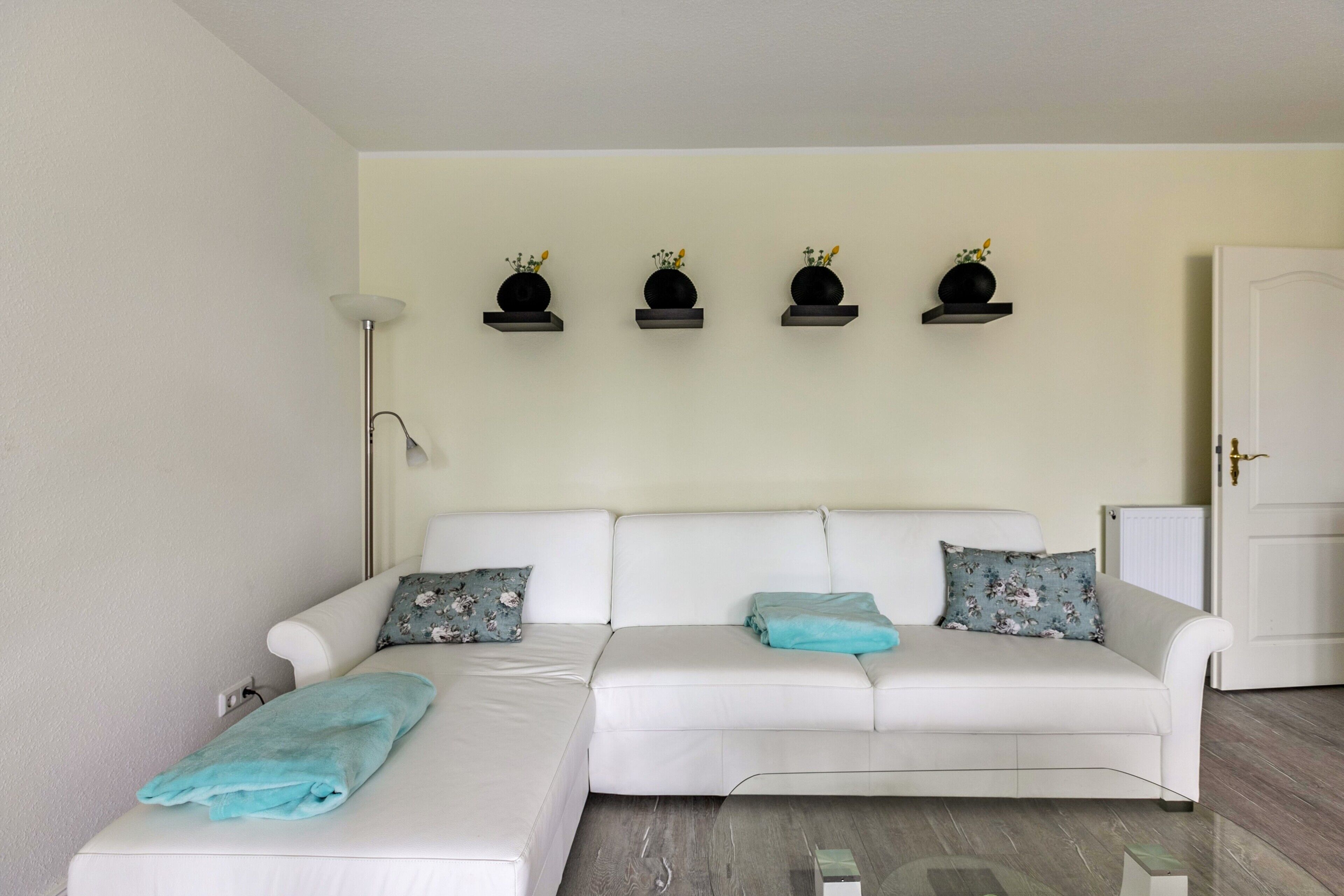A modern living room with a white sofa, a floor lamp, and three black shelves with flowers.