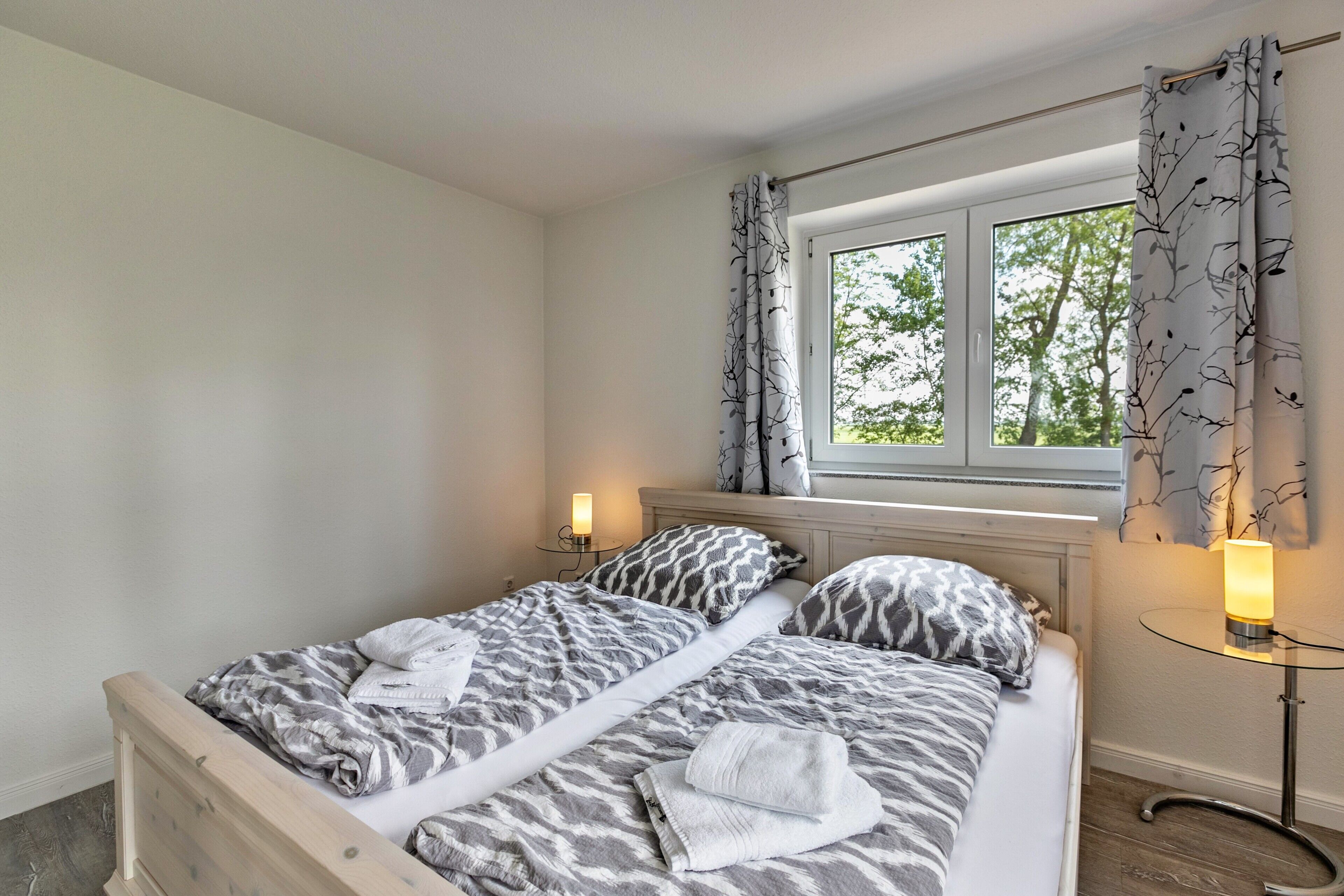 Un lit double avec une couvre-lit à motifs zébrés, deux tables de chevet avec des lampes, une fenêtre avec des rideaux et une vue sur les arbres à l’extérieur.