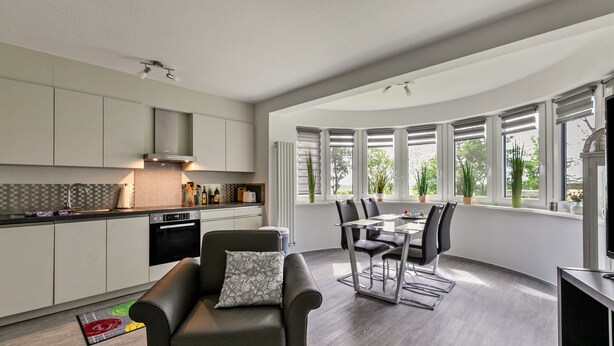 Una cocina moderna con armarios blancos, una zona de comedor con mesa y sillas, y ventanas amplias con persianas.