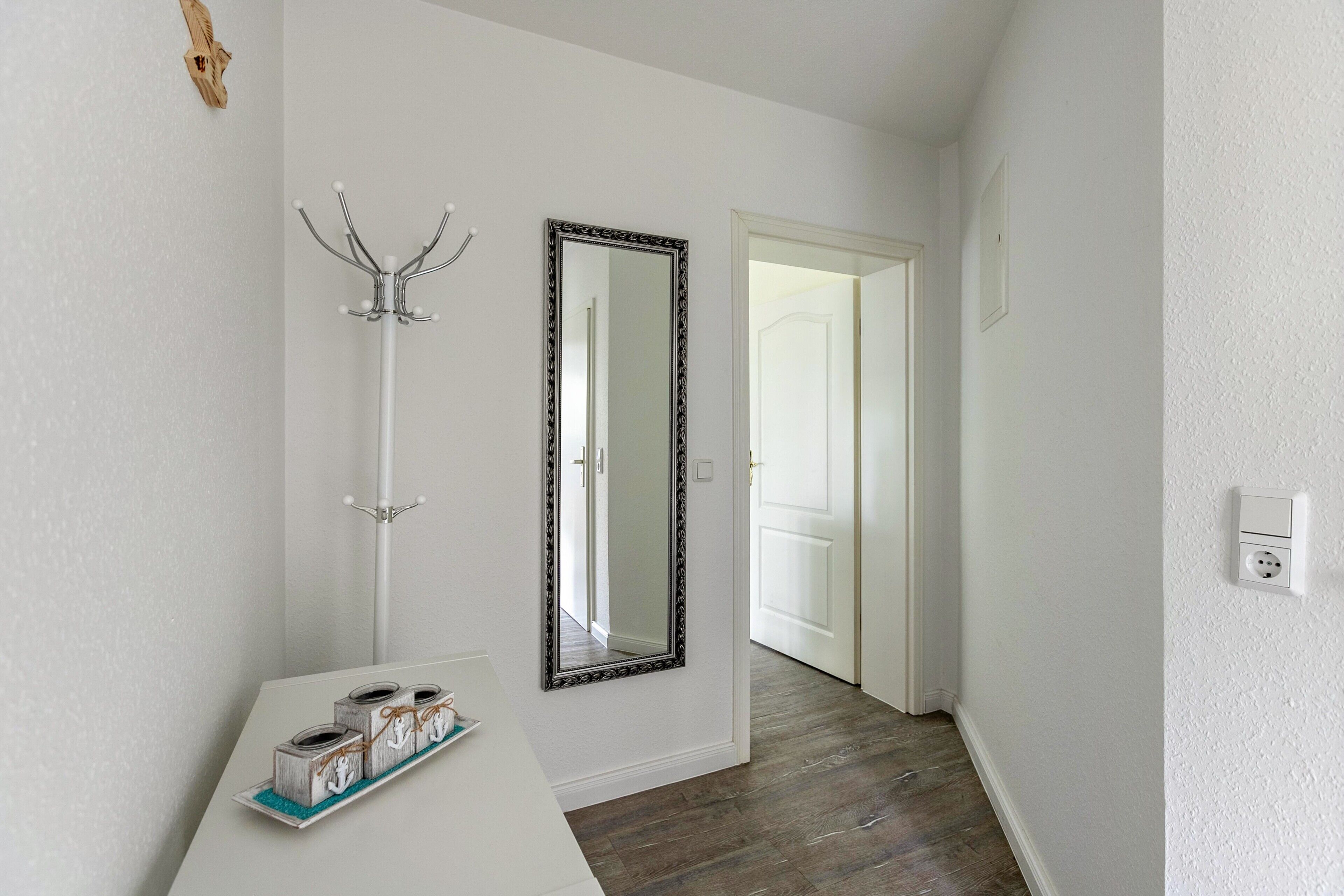 A hallway with a white wall, a large mirror, a coat rack, and a wooden floor.