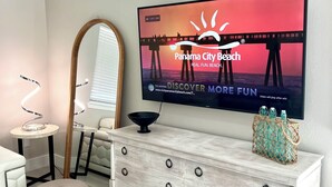 Een moderne woonkamer met een grote flatscreen-tv waarop een advertentie voor een strand te zien is, een witte bank, een ronde salontafel, een spiegel met houten lijst en een lichtkleurige houten kast met lades.