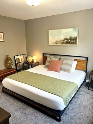 A neatly made bed with a green throw blanket, a wooden headboard, and multiple pillows. There is a bedside table with a lamp, a vase, and a framed picture. A large abstract painting hangs on the wall above the bed. - Grand Lagoon Vista -air hockey- Dreamy Day (Panama City Beach)