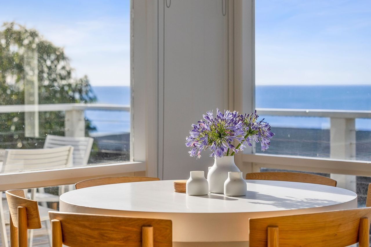 Une table ronde ornée d’un vase de fleurs violettes, de bougeoirs blancs et offrant une vue sur l’océan depuis un balcon.