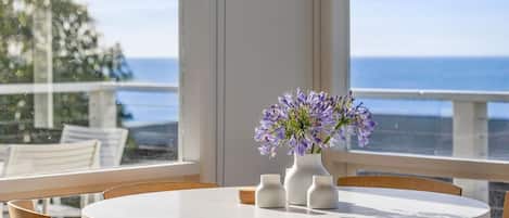 Une table ronde ornée d’un vase de fleurs violettes, de bougeoirs blancs et offrant une vue sur l’océan depuis un balcon.