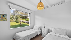 Ein Schlafzimmer mit zwei Einzelbetten, einem Nachttisch, einer Lampe, einem Fenster mit Blick ins Freie und einer Pendelleuchte.
