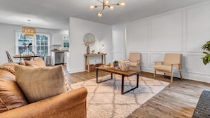 Een moderne woonkamer met een leren bank, een salontafel en een beige fauteuil. Achtergrond is een keuken met witte kasten en een eethoek met een tafel en stoelen.