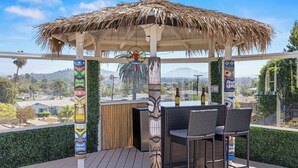 Un espace extérieur abrité avec des tables et des chaises, un toit de chaume, des totems tiki et une vue sur la ville et les montagnes.