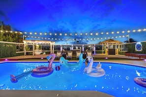Uma piscina com brinquedos infláveis, área de estar coberta e luzes de corda.