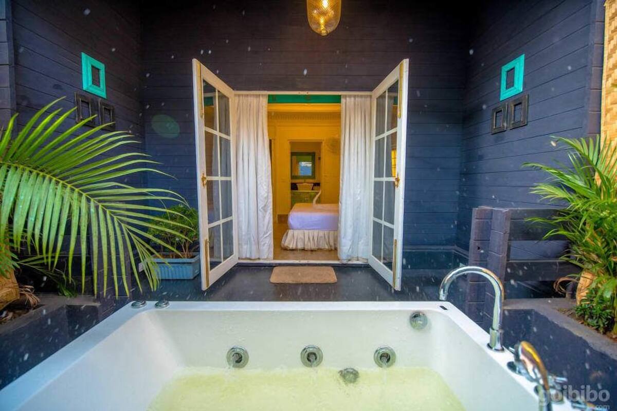 Quarto com uma banheira grande, cama com colcha branca e vista para outro espaço através de uma porta aberta.
