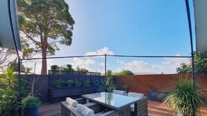 An outdoor patio with a glass-top table and wicker furniture, surrounded by potted plants and a wooden deck.
