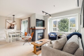 A modern living room with a fireplace, a dining area, and a large window with a view of trees.