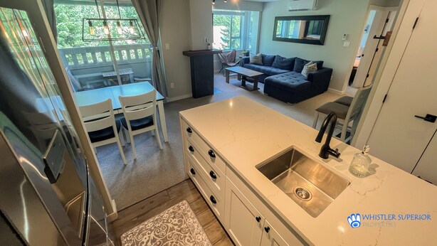 A modern kitchen with a large island, a dining area with a table and chairs, a dark-colored sofa, and a view of greenery outside. - Renovated Whistler Village Condo with Stunning Blackcomb Views (Whistler)