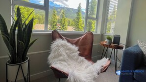 A room with a large window offering a mountain view, a brown leather chair with white fur trim, a potted plant, and a small table with a speaker. - Renovated Whistler Village Condo with Stunning Blackcomb Views (Whistler)