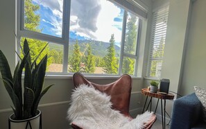 A room with a large window offering a mountain view, a brown leather chair with white fur trim, a potted plant, and a small table with a speaker.